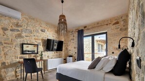 A bedroom with a stone wall, a bed, a desk, a chair, a TV, and a window with a view. - ALAGNI CRETAN RESORT (Alagni)