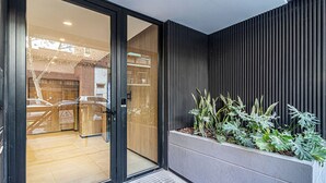 Ein moderner Gebäudeeingang mit Glastür, Metallgitter und einem Pflanzgefäß mit Pflanzen.