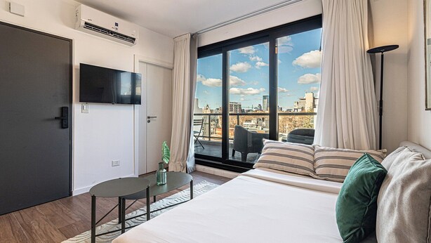 Ein modernes Hotelzimmer mit einem großen Bett, einem an der Wand befestigten Fernseher, einem kleinen Tisch mit einer Pflanze und Blick auf die Stadt durch große Fenster.