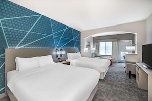 Habitación de hotel con dos camas, un escritorio con televisor, una silla y un baño visible a través de un arco.