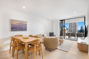 A modern living room with a dining area, a sofa, a coffee table, and a view of the cityscape through large windows. - The Coastal Luxe Haven - Coolangatta (Gold Coast)
