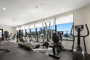 A modern gym with cardio machines, weightlifting equipment, and large windows offering a city view. - The Coastal Luxe Haven - Coolangatta (Gold Coast)