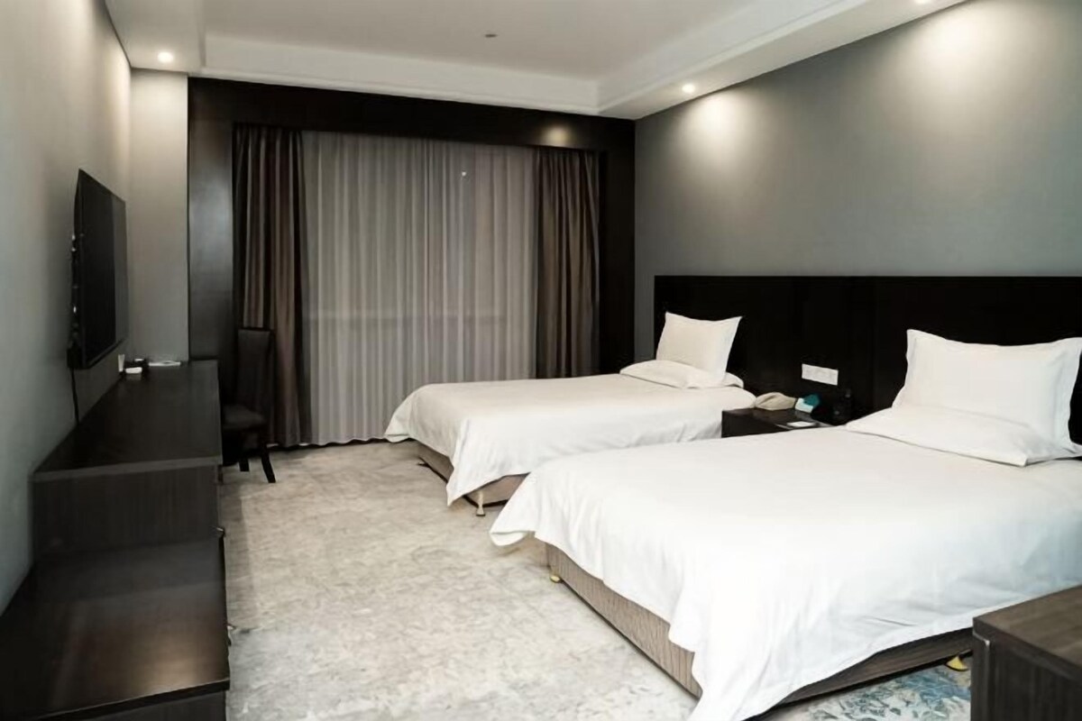 A hotel room with two beds, a desk, a TV, and a large window with curtains.