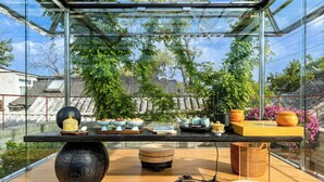 Um interior moderno com estrutura de vidro, uma mesa de madeira com diversos objetos e vista para o verde lá fora.