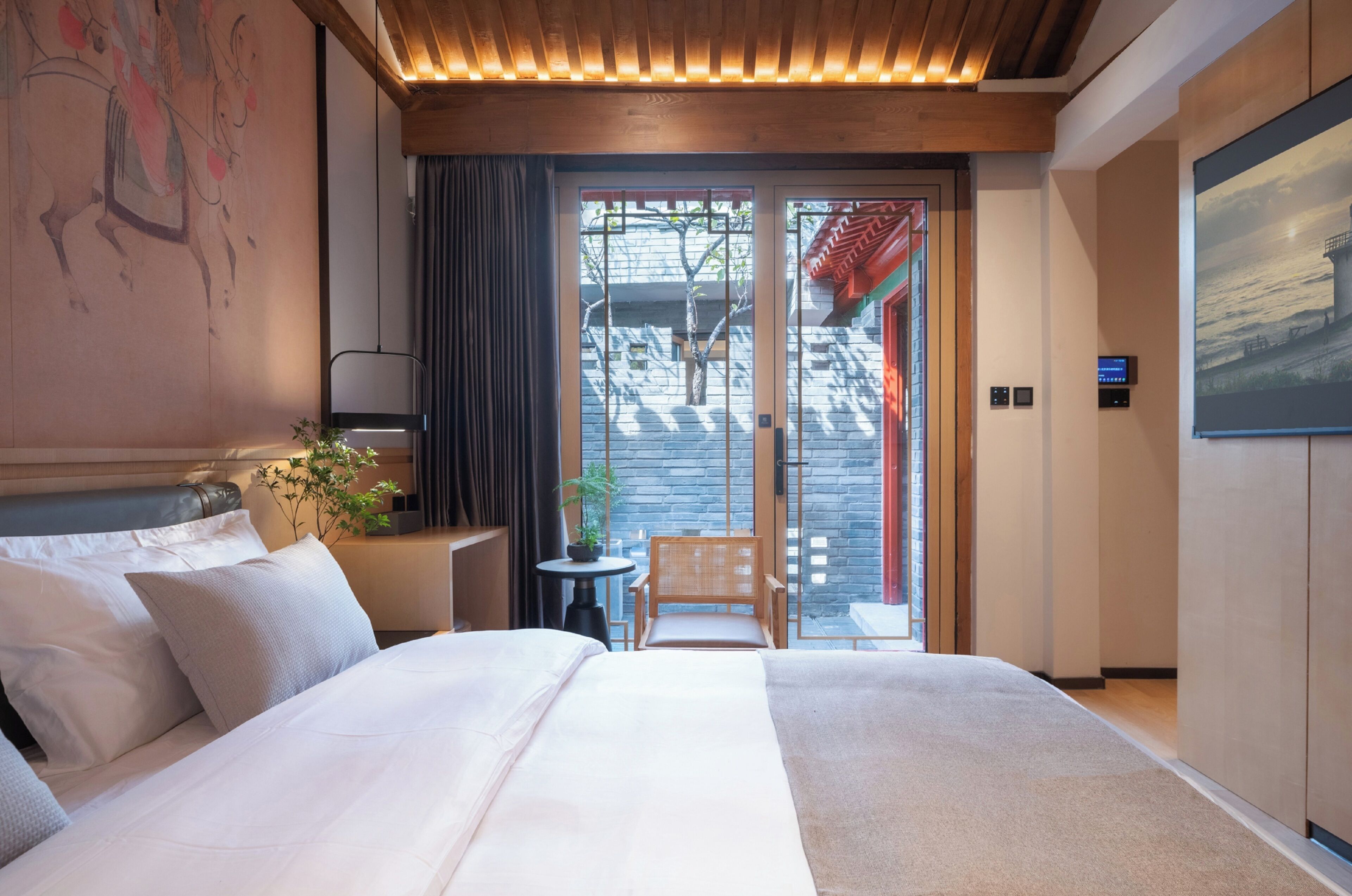 A hotel room with a large bed, a small table with a chair, a potted plant, and a view of the outside through a sliding door.