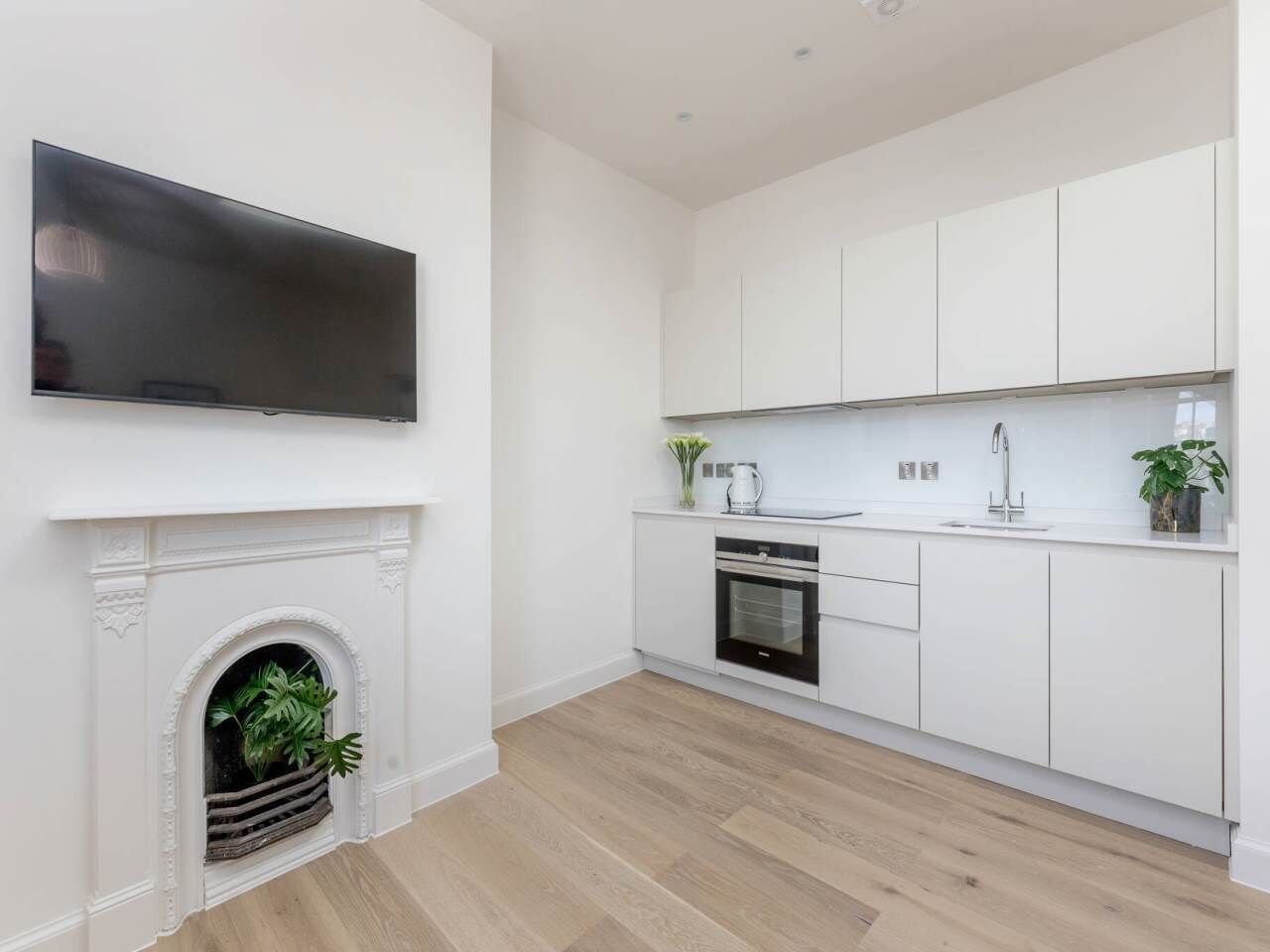 A modern kitchen with white cabinets, a flat-screen TV, a fireplace, and a small plant.