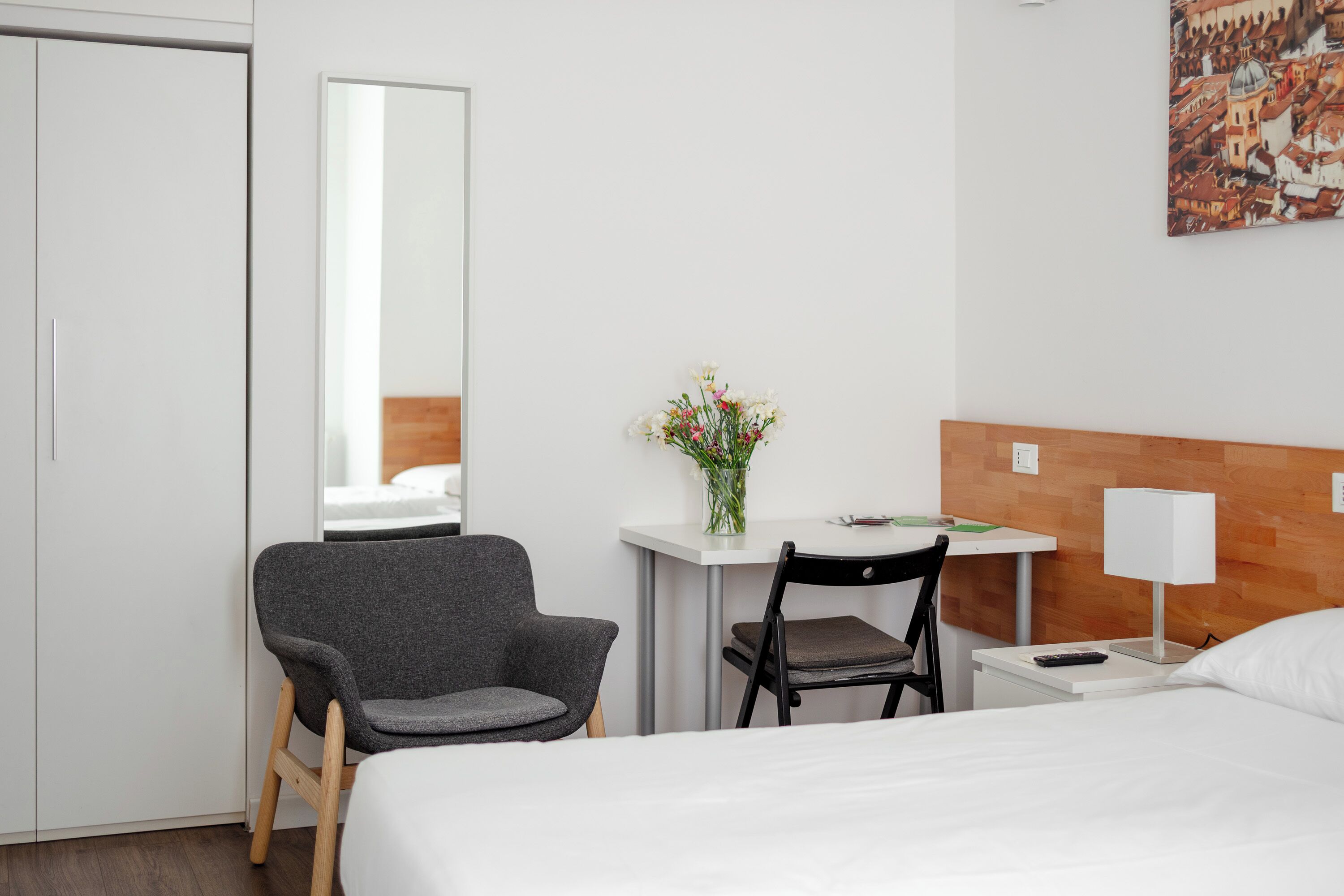 A hotel room with a bed, a desk with a chair, a small table with a lamp, a mirror, and a wall-mounted painting.