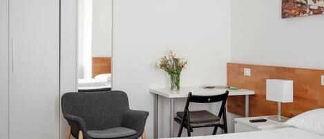 A hotel room with a bed, a desk with a chair, a small table with a lamp, a mirror, and a wall-mounted painting.
