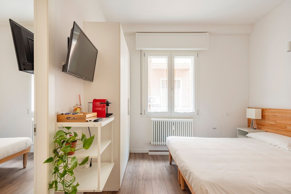 Ein Schlafzimmer mit einem Bett, einem an der Wand befestigten Fernseher, einem Heizkörper und einem Fenster mit Jalousien.