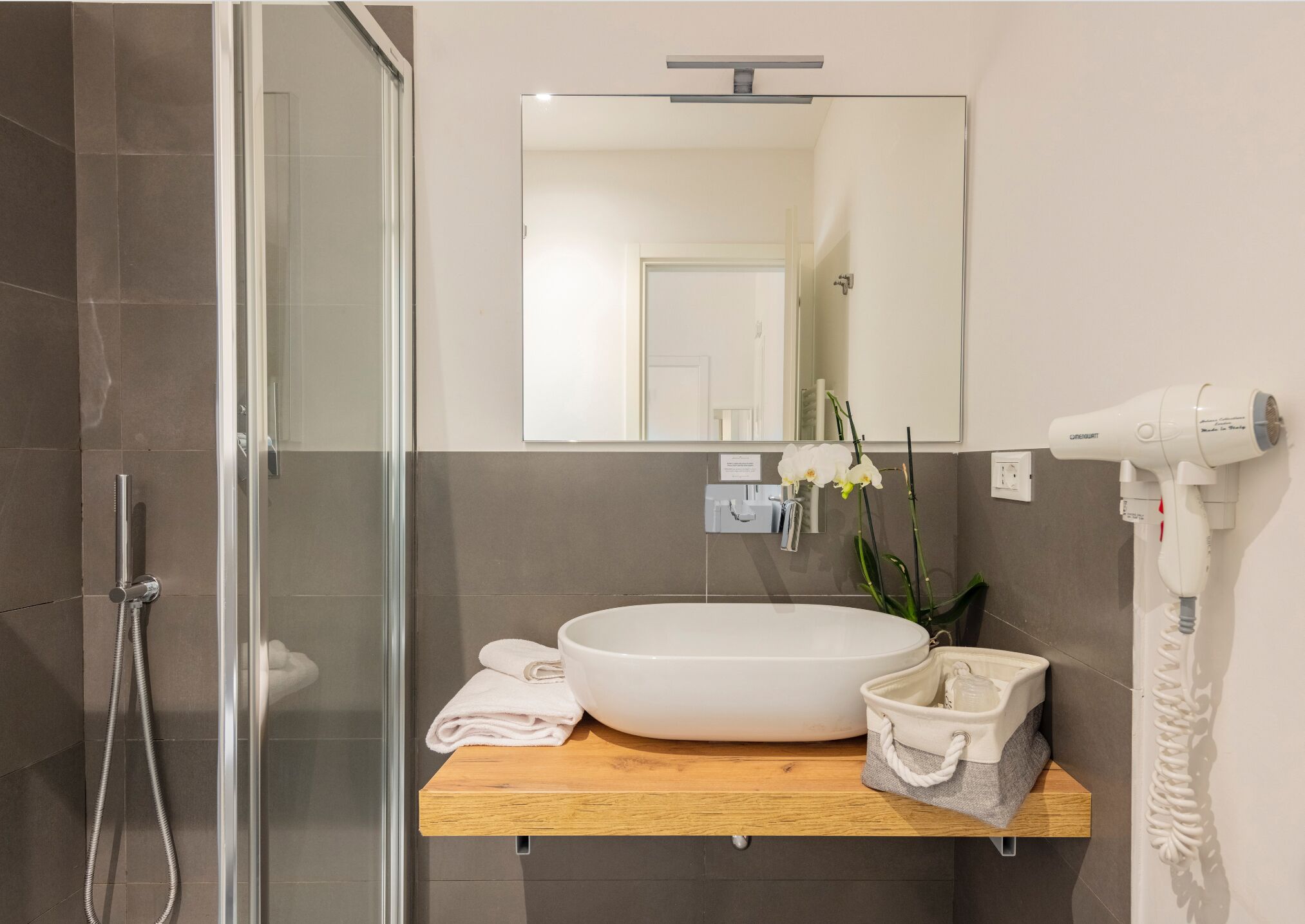 Un bany amb mampara de vidre, un lavabo blanc, un mirall, un assecador i una petita planta.