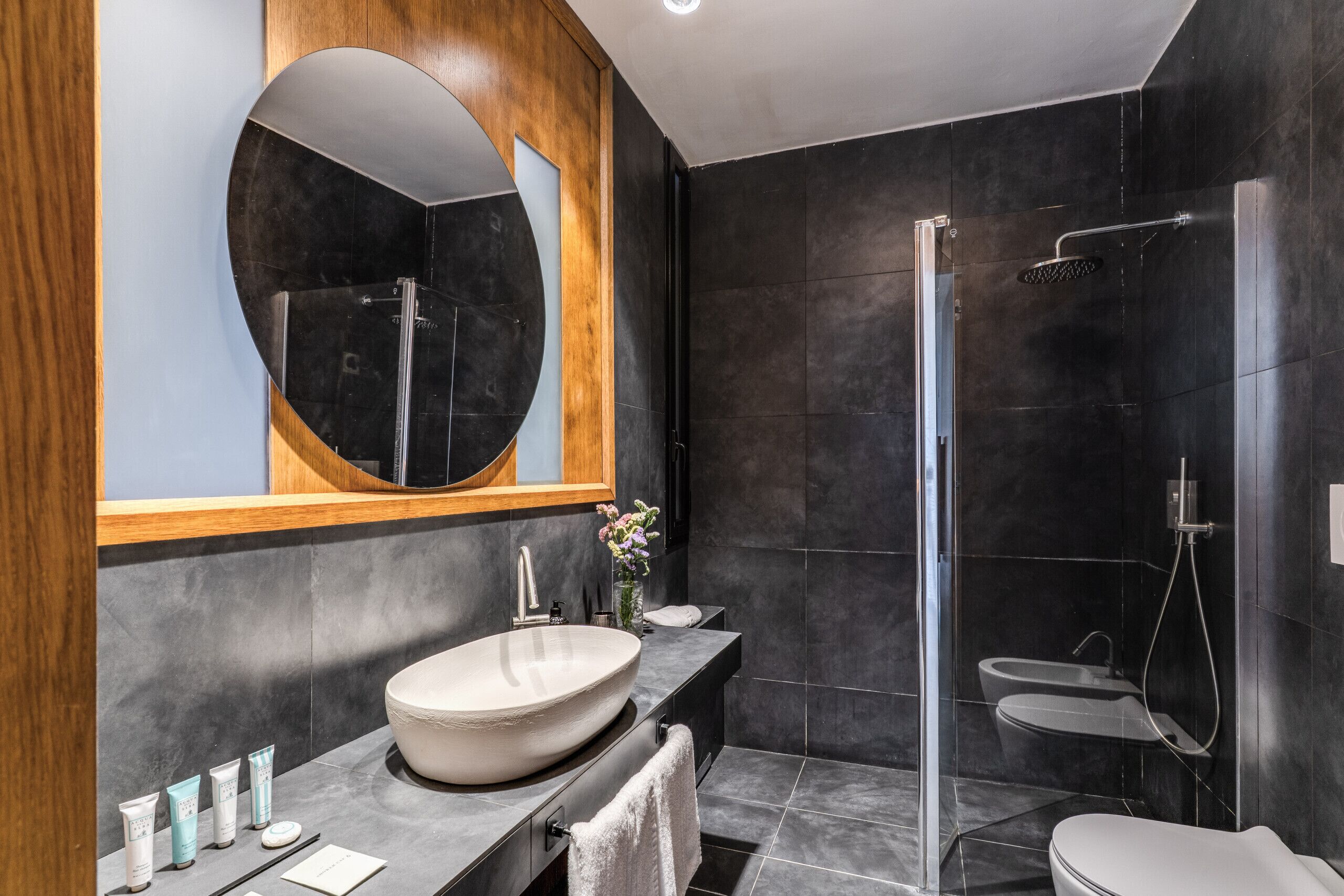 Een moderne badkamer met een grote ronde spiegel, een wastafel op voet, een douche met glazen wanden en een toilet.