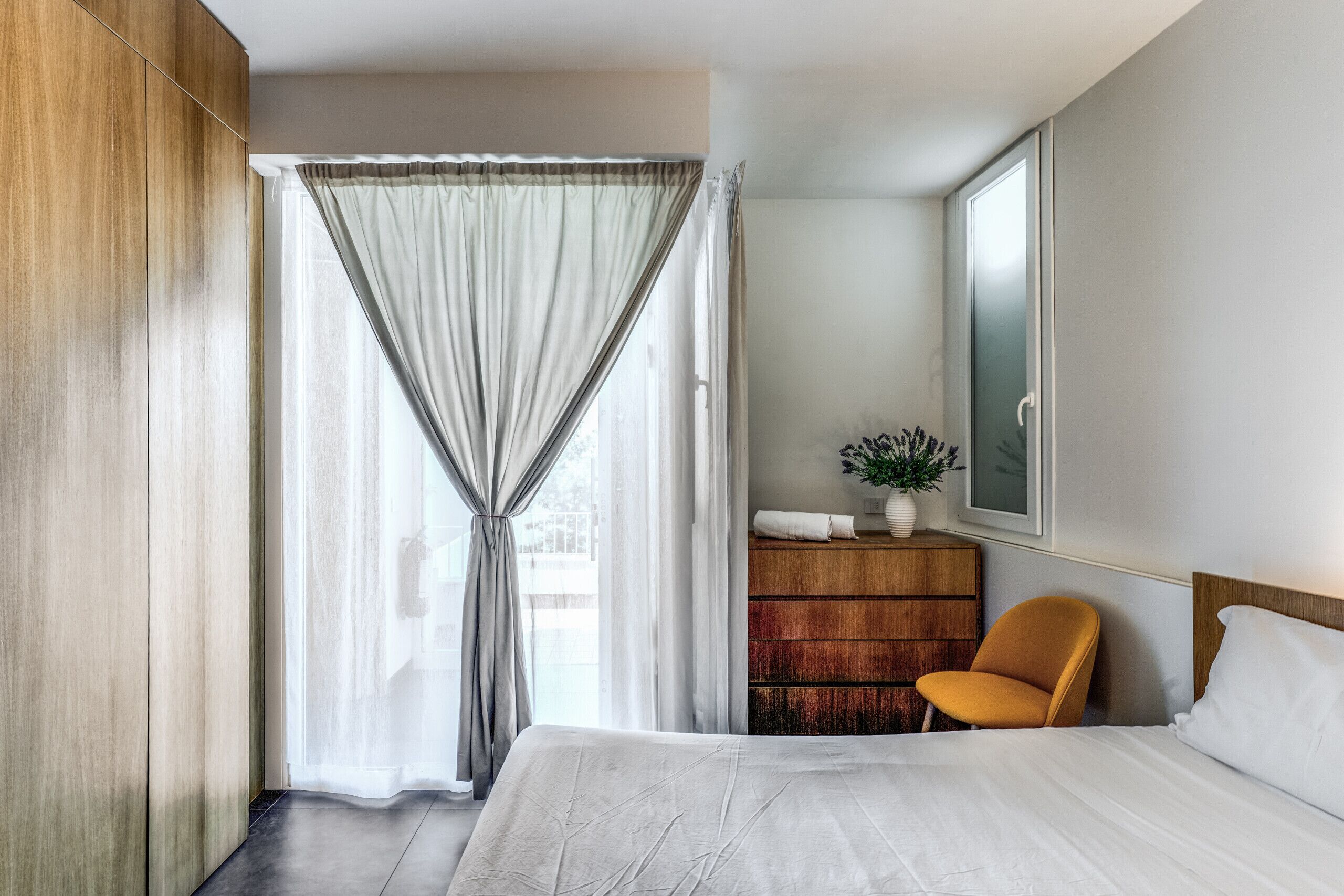 Une chambre d’hôtel avec un lit, une armoire en bois, une commode en bois, une chaise et une fenêtre avec des rideaux.