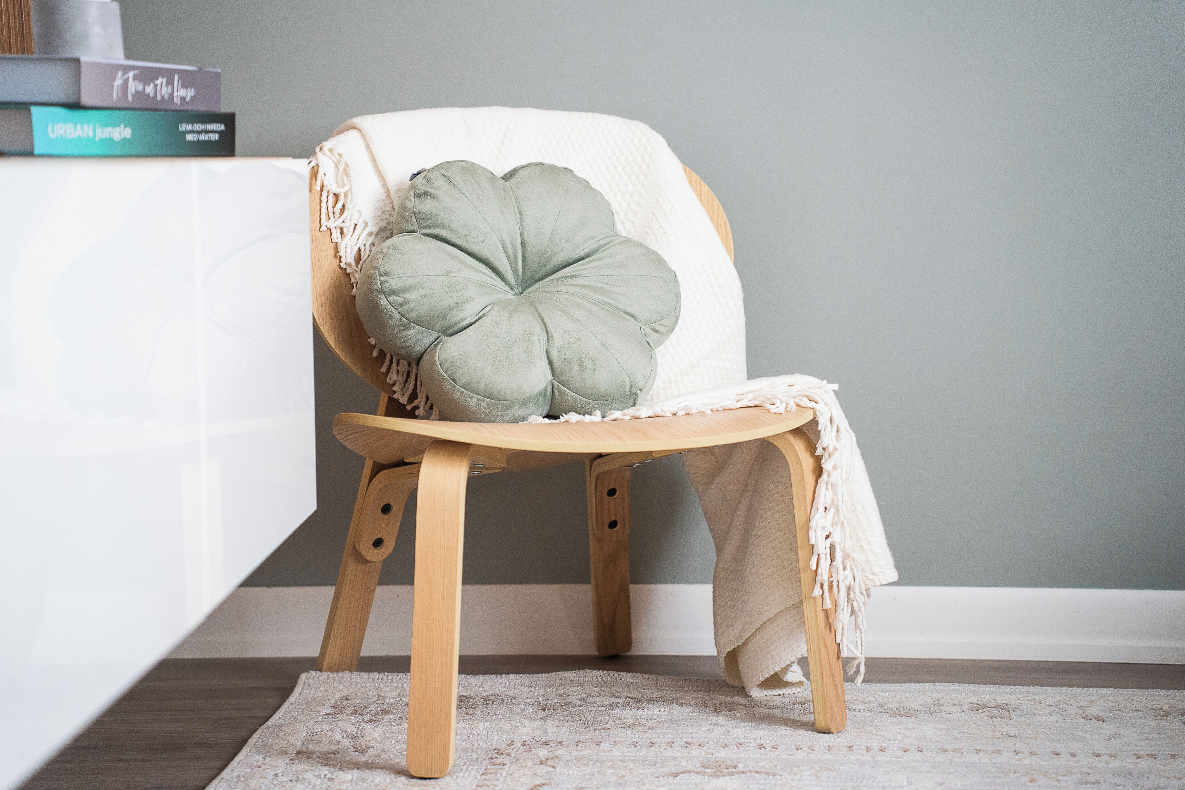 Une chaise en bois avec un coussin, une couverture blanche et une petite pile de livres posée sur une surface blanche.