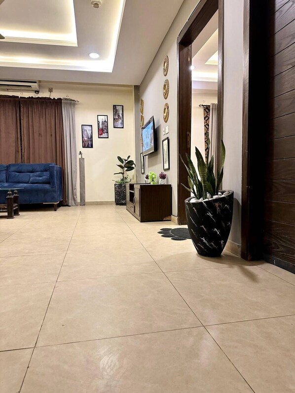 A spacious living room with a blue sofa, a flat-screen TV, decorative wall art, and a large potted plant.