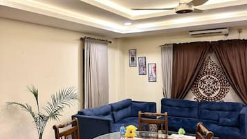 Sala de estar com mesa de jantar de vidro, sofás azuis, ventilador de teto e um objeto decorativo na parede.