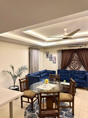 A living room with a glass dining table, blue sofas, a ceiling fan, and a decorative wall piece.