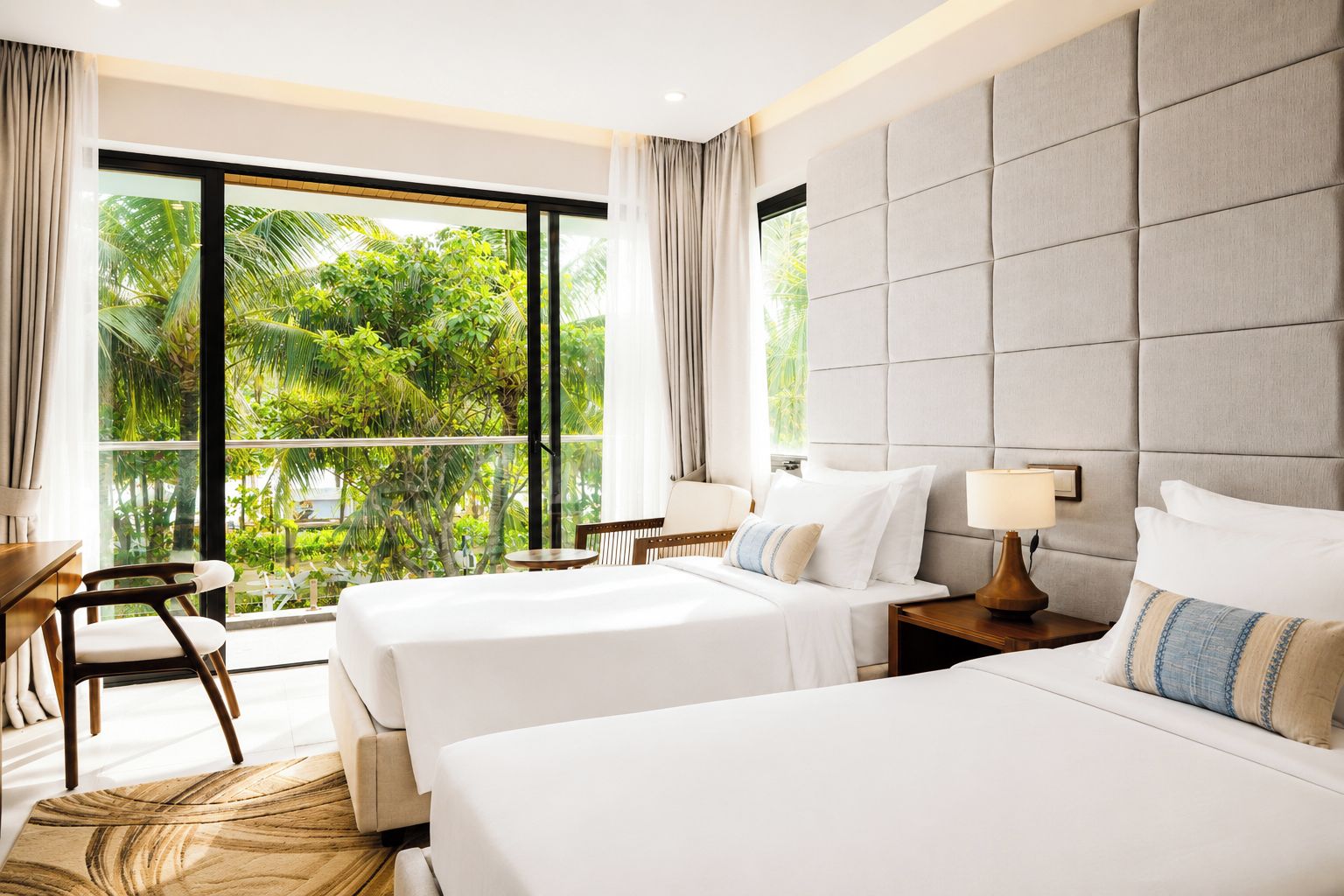 A hotel room with two beds, a desk, and a chair. There is a large window with a view of greenery outside.