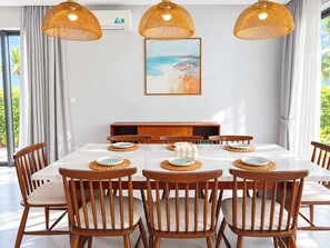 A dining area with a white table, wooden chairs, woven placemats, and a beach-themed painting on the wall. - Sonasea Villas Collection Long Beach Phu Quoc (Phu Quoc)