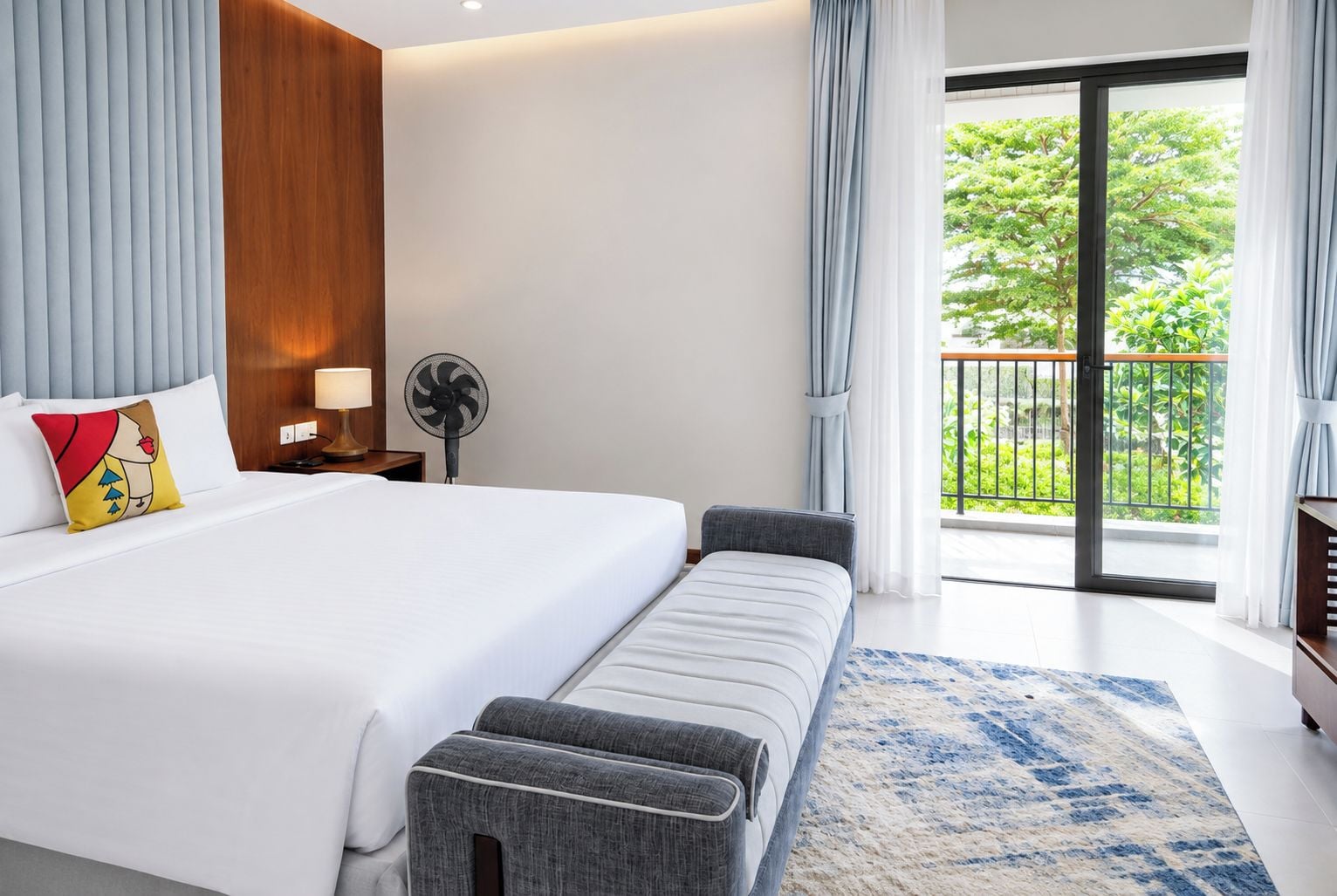 Habitación de hotel moderna con una cama grande, un banco, un ventilador y vistas a zonas verdes a través del balcón.