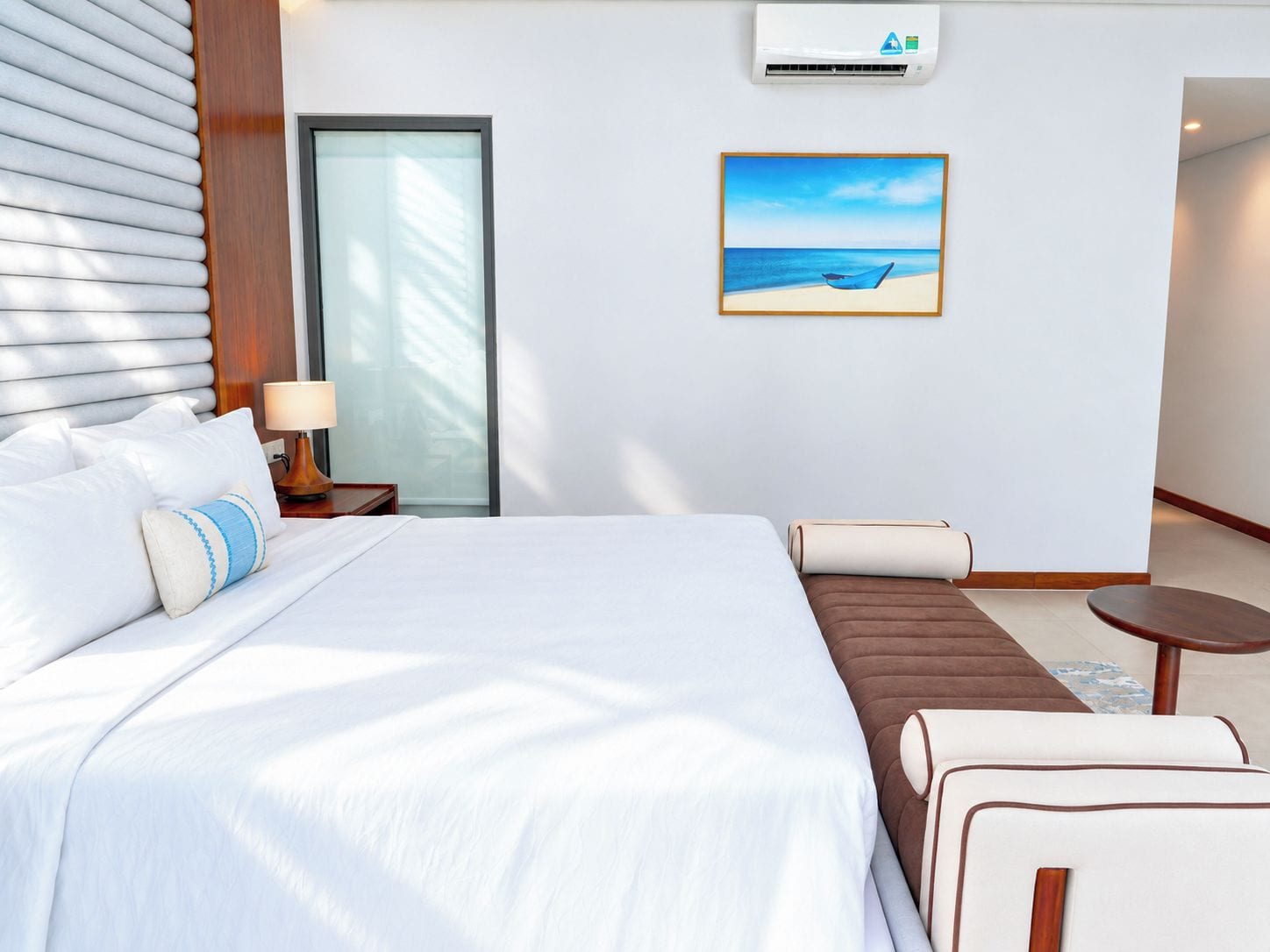 Ein Hotelzimmer mit einem großen Bett, einem Nachttisch mit Lampe, einem Gemälde mit Strandmotiv und Blick auf den Ozean durch ein Fenster.