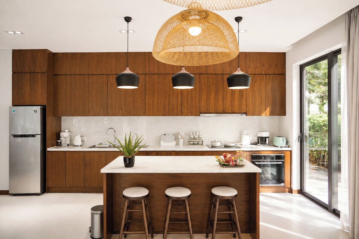 A modern kitchen with wooden cabinets, a stainless steel refrigerator, a central island with stools, pendant lighting, and a view of greenery outside.