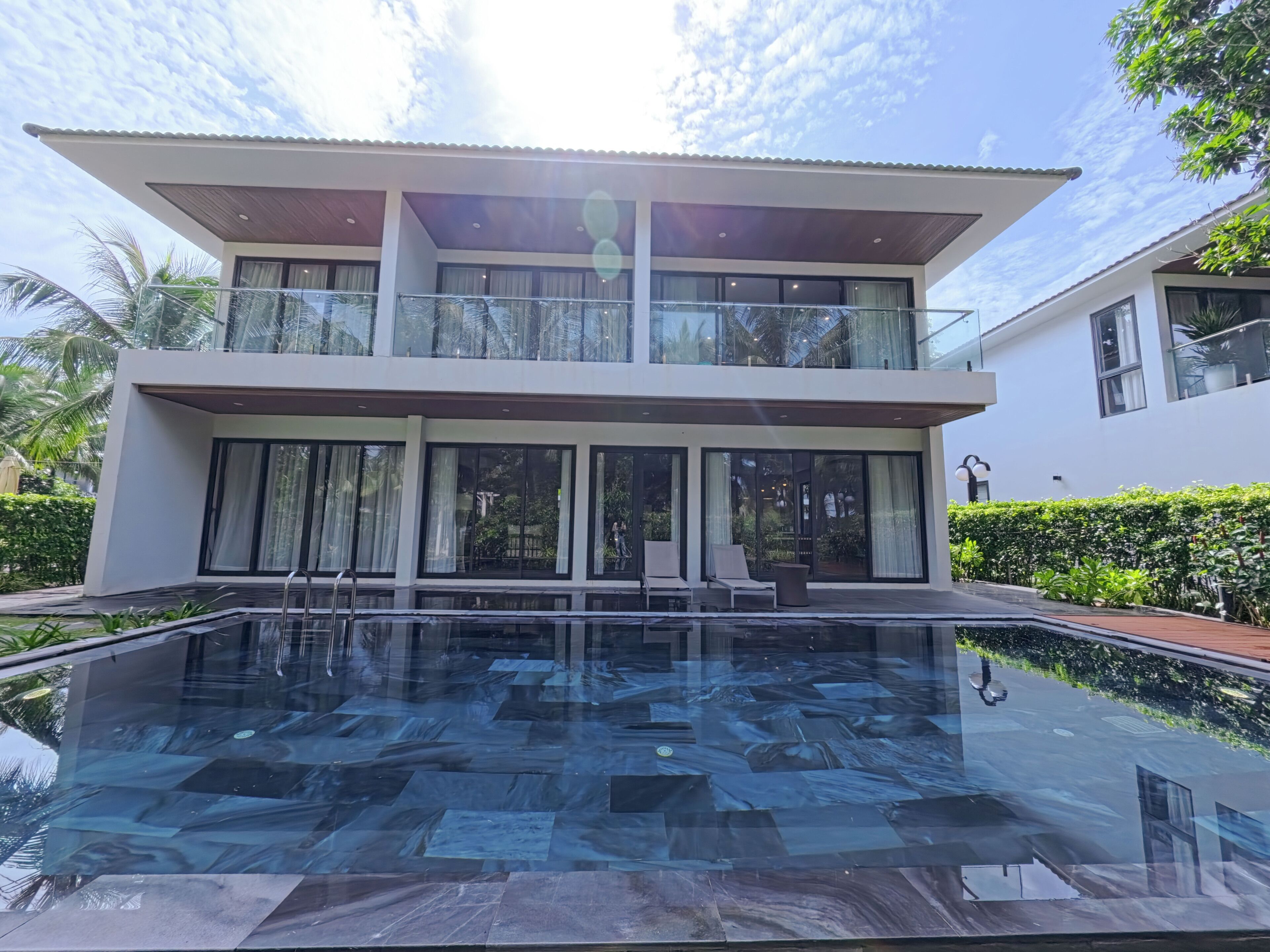 Una casa moderna de dos pisos con balcón de vidrio, piscina y exuberante vegetación.