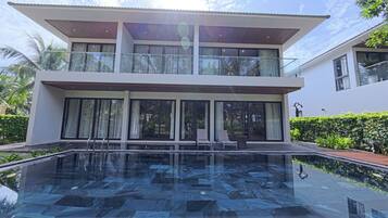 Una casa moderna de dos pisos con balcón de vidrio, piscina y exuberante vegetación.