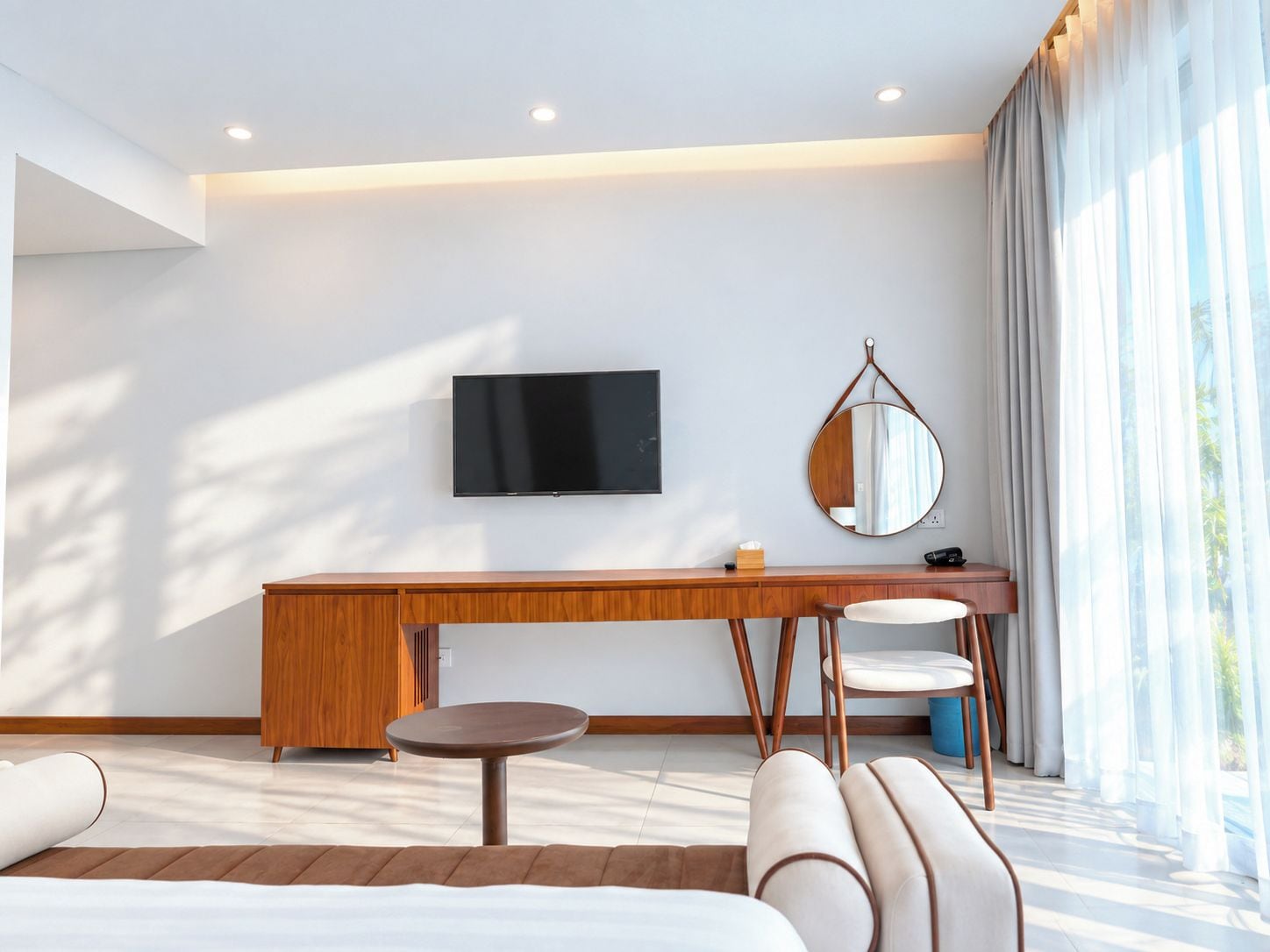 Habitación de hotel moderna con televisor de pantalla plana, espejo redondo, escritorio de madera, mesa redonda y sofá.