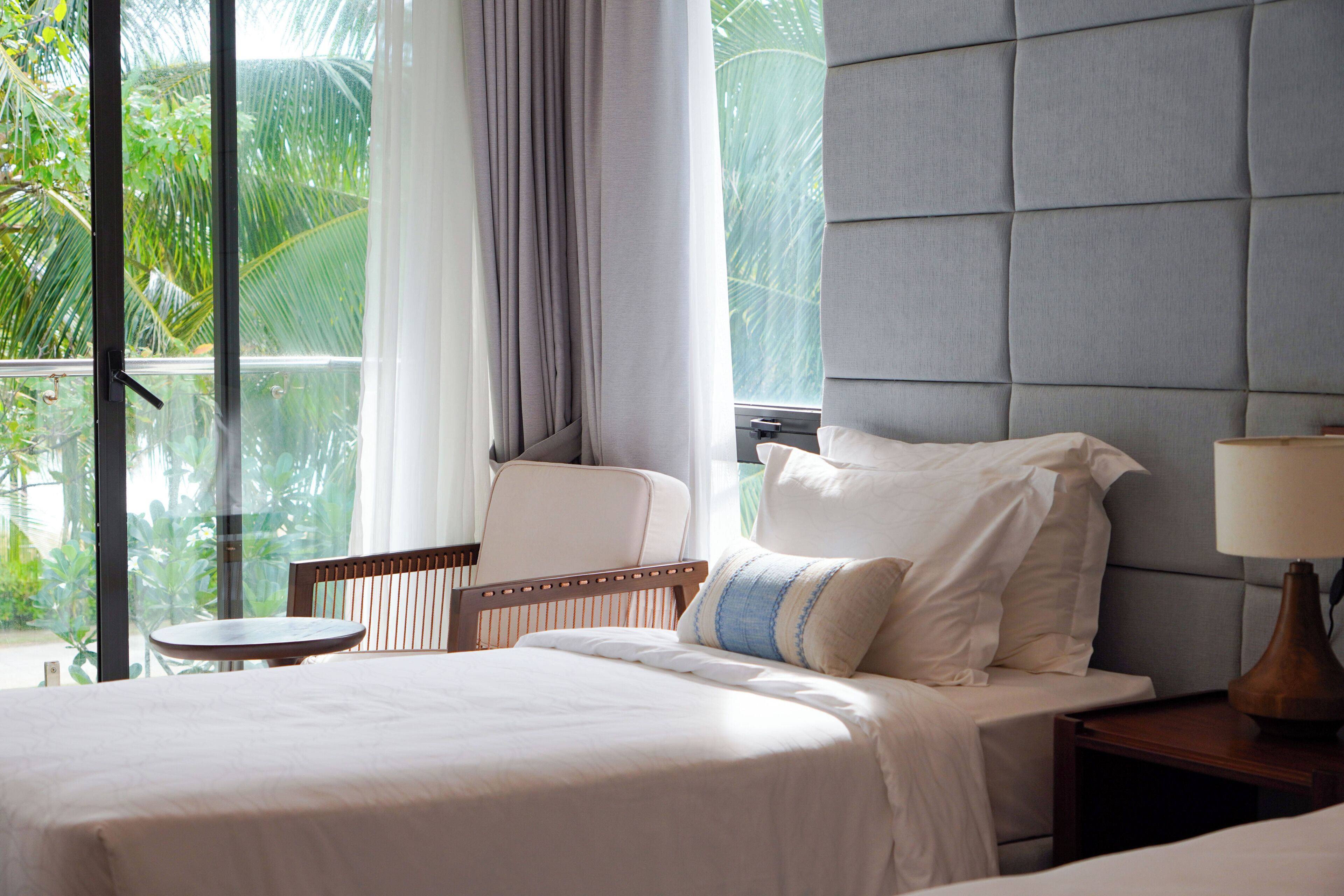 A hotel room with a bed, a chair, a nightstand, a lamp, and a view of greenery through the window.