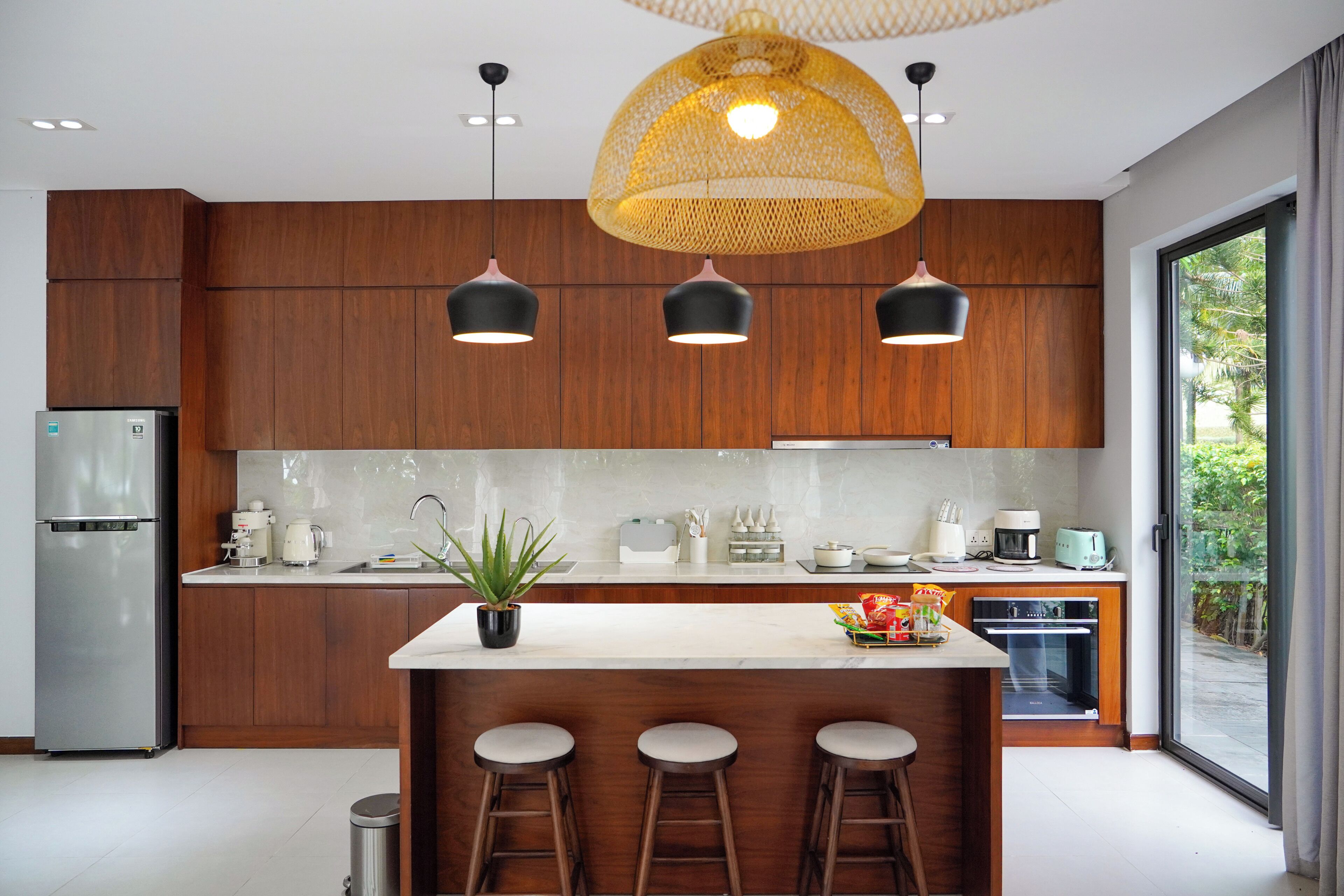 A modern kitchen with a central island, bar stools, a refrigerator, and a large window with a view of greenery.