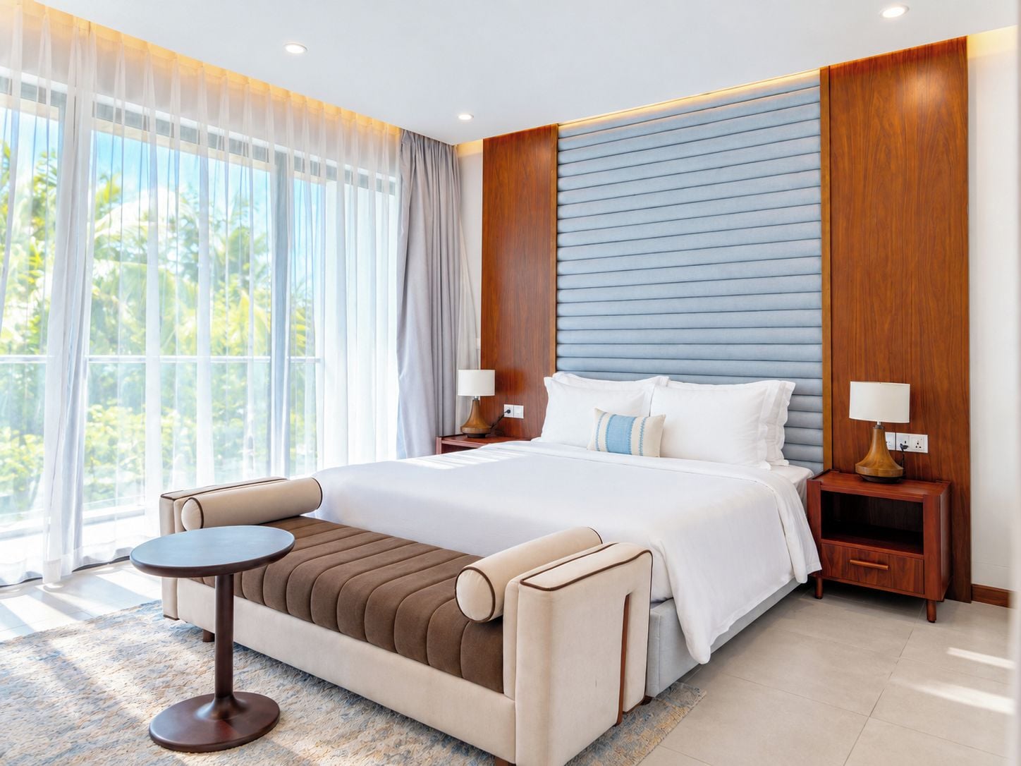 A modern hotel room with a large bed, a sofa bed, a small round table, and a nightstand with lamps.