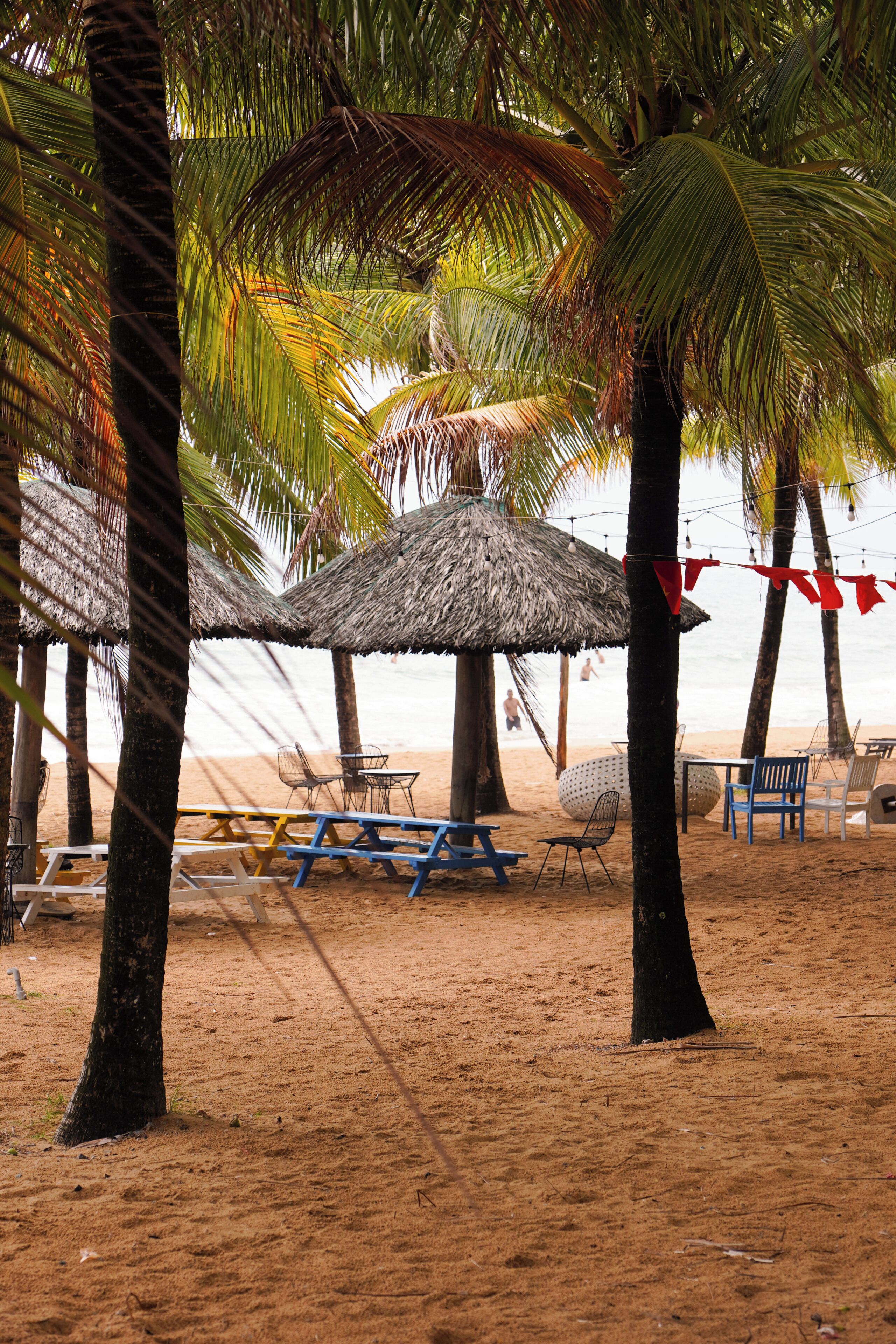 Escena de playa con sombrillas de paja, mesas de picnic y sillas.