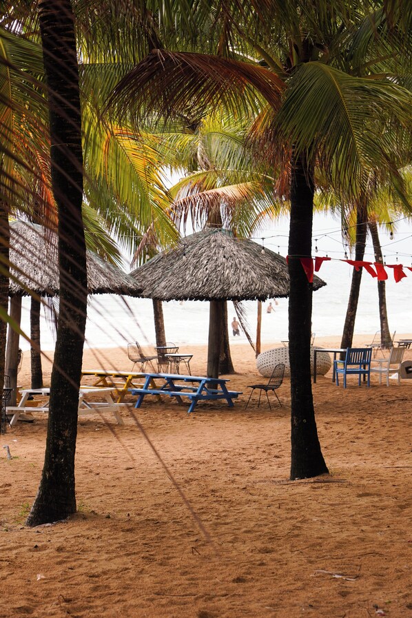 Escena de playa con sombrillas de paja, mesas de picnic y sillas.