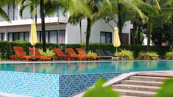 Zona de piscina con sillas de descanso y sombrillas, rodeada de palmeras y un edificio con grandes ventanales.
