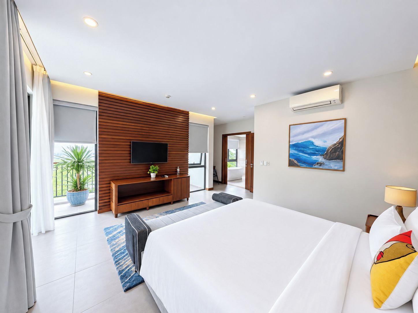 Habitación de hotel moderna con una cama grande, un televisor de pantalla plana montado en una pared de paneles de madera, un cuadro de una escena marina y un balcón con una planta en maceta.
