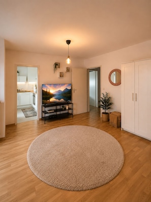 Een ruime woonkamer met een groot rond vloerkleed, een flatscreen televisie aan de muur, een plafondlamp en een potplant.