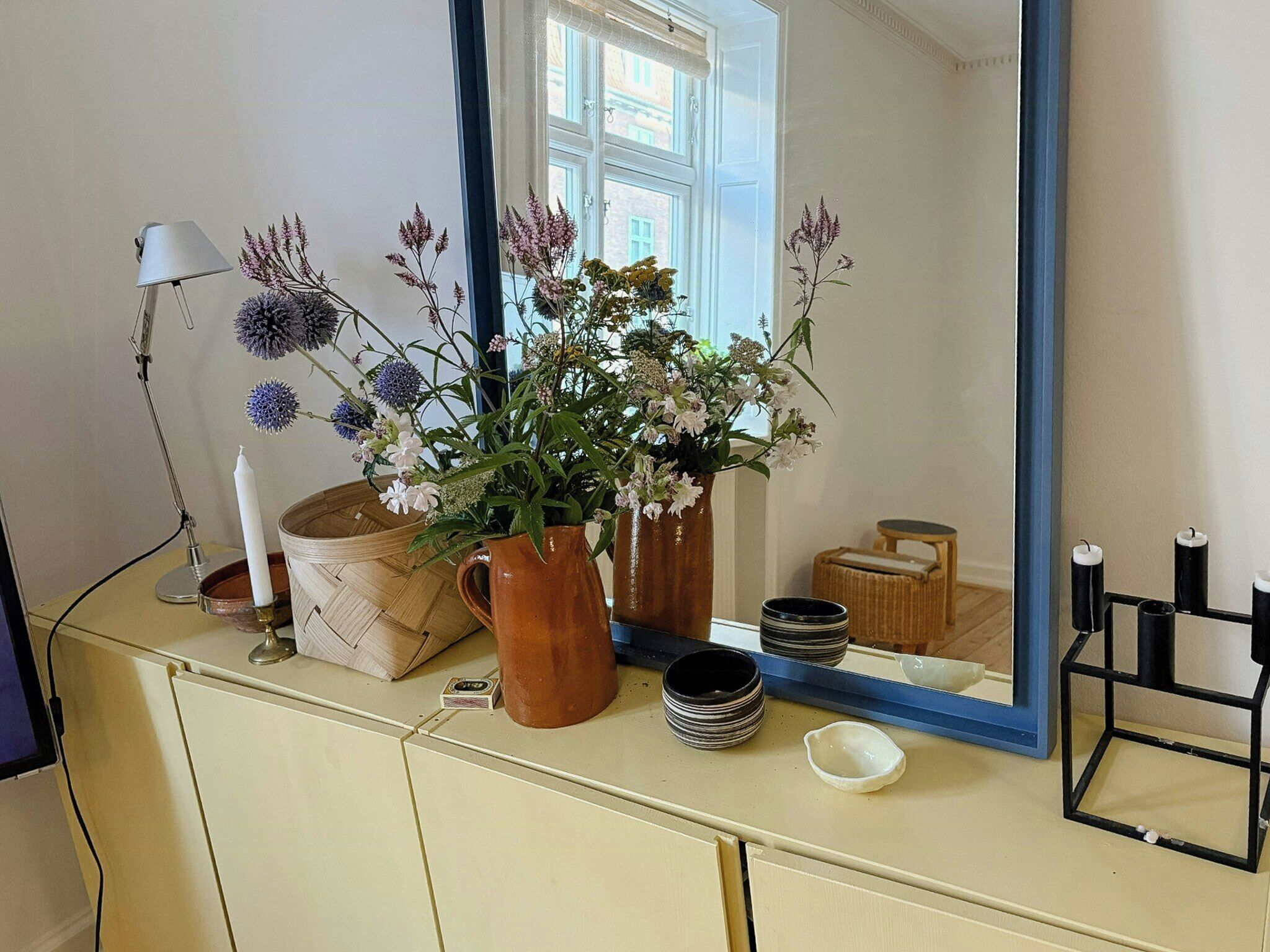 Ett rum med en spegel som reflekterar ett fönster, en lampa, en vas med blommor och ett litet bord med ljus.