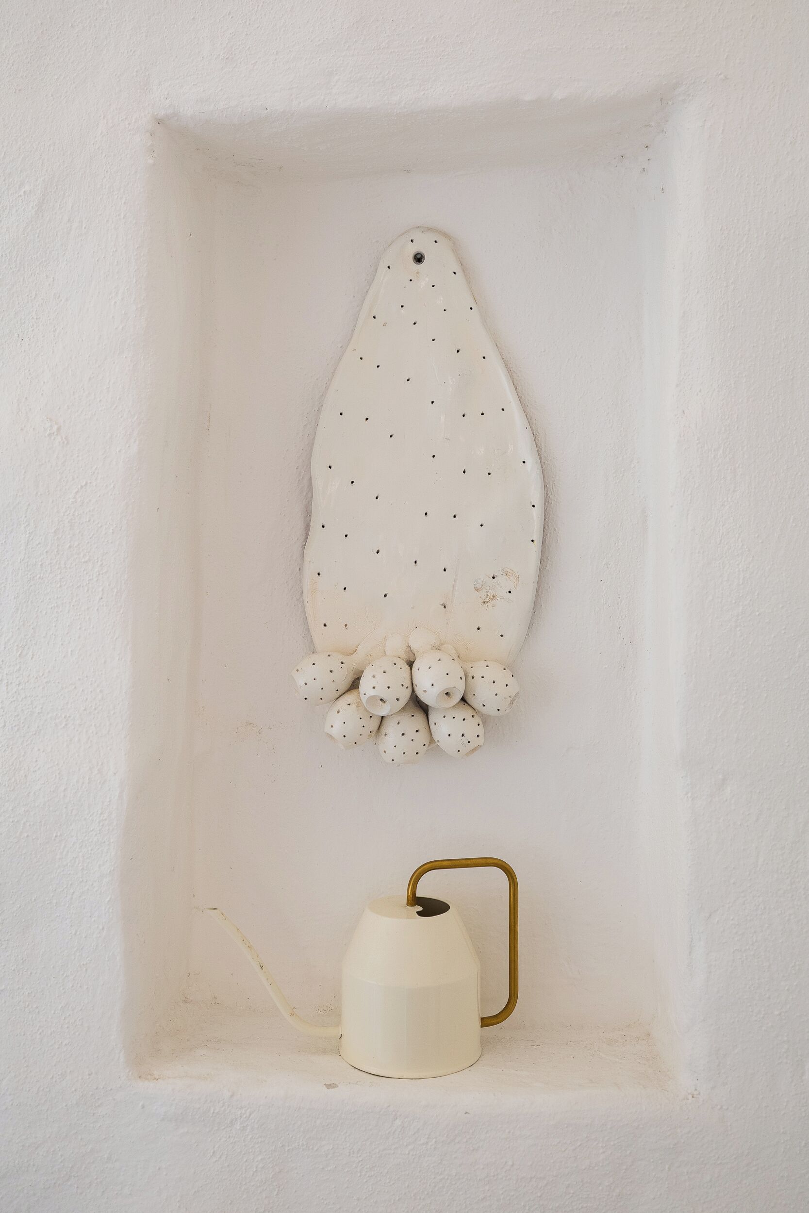 A white wall with a recessed area containing a white ceramic object with holes and a white ceramic mug with a gold handle.
