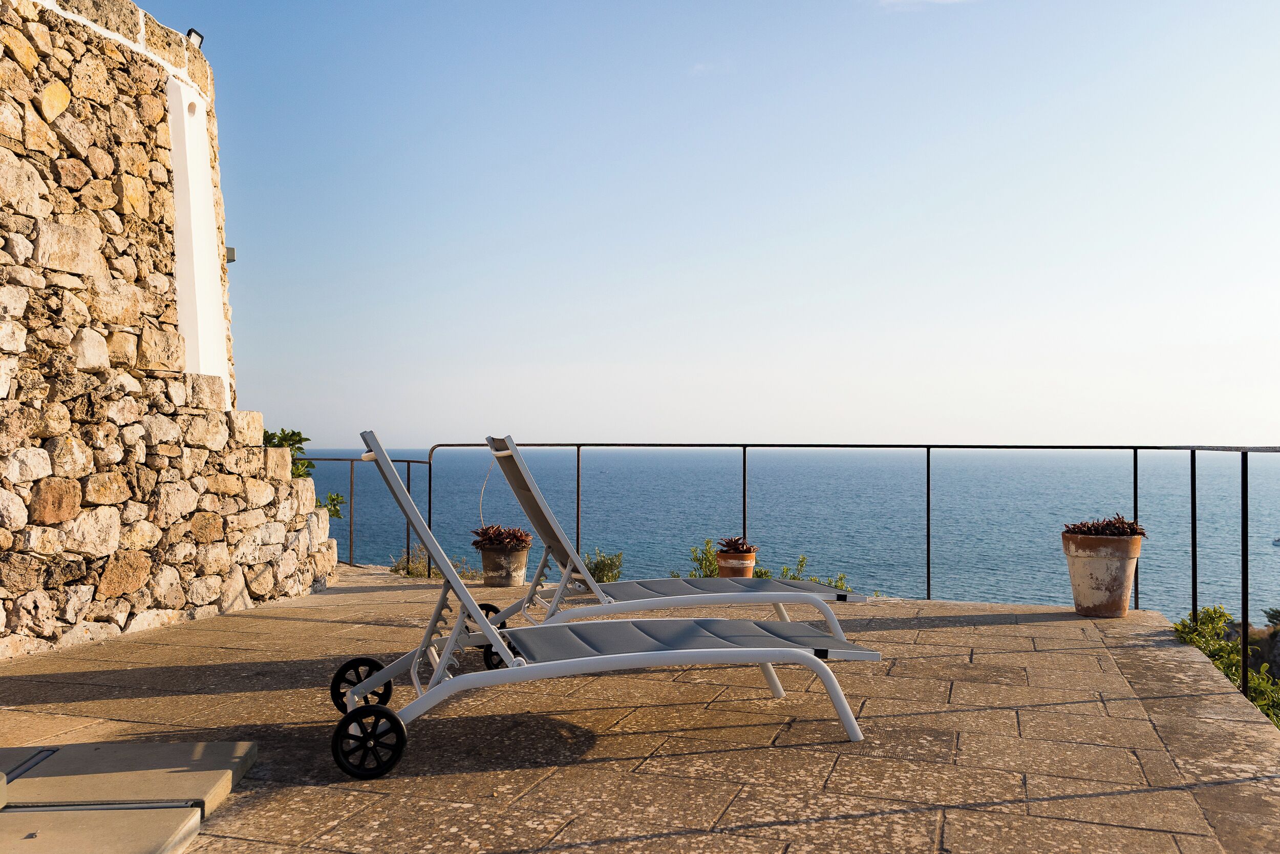 Un balcón con pared de piedra, una silla de metal y plantas en macetas, con vista al mar.