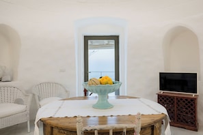 A room with a round wooden table, a white wicker chair, a teal bowl with fruit, a television on a wooden stand, and a white door with a view of the sky. - TRULLO EXCLUSIVE (Torre Vado)