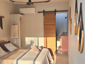 Een slaapkamer met een bed, een plafondventilator, een airconditioning en een macramé wanddecoratie, met een schuifdeur naar een badkamer.
