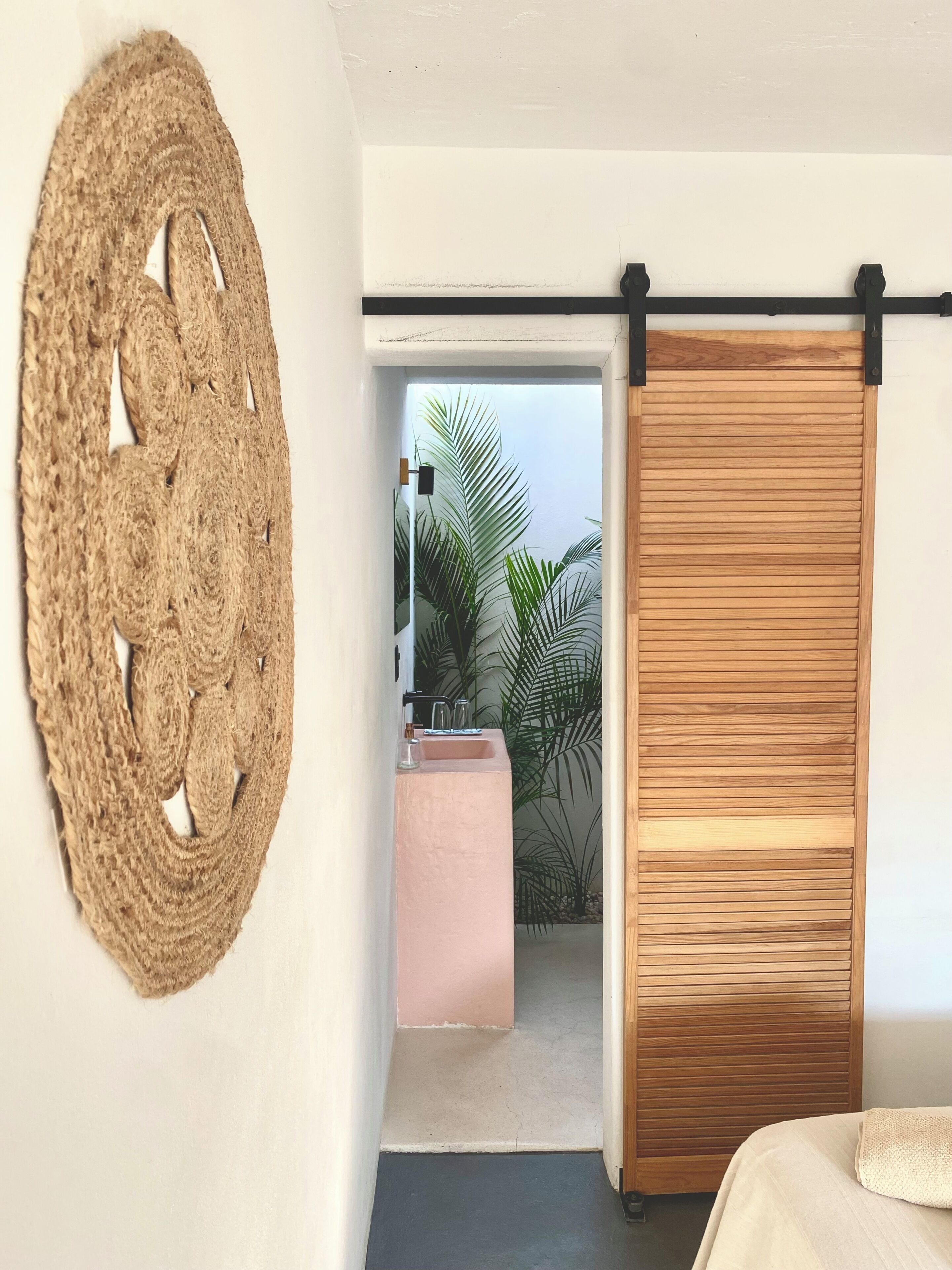 A room with a bed, a wall-mounted woven artwork, a sliding wooden door, and a view of a balcony with plants.