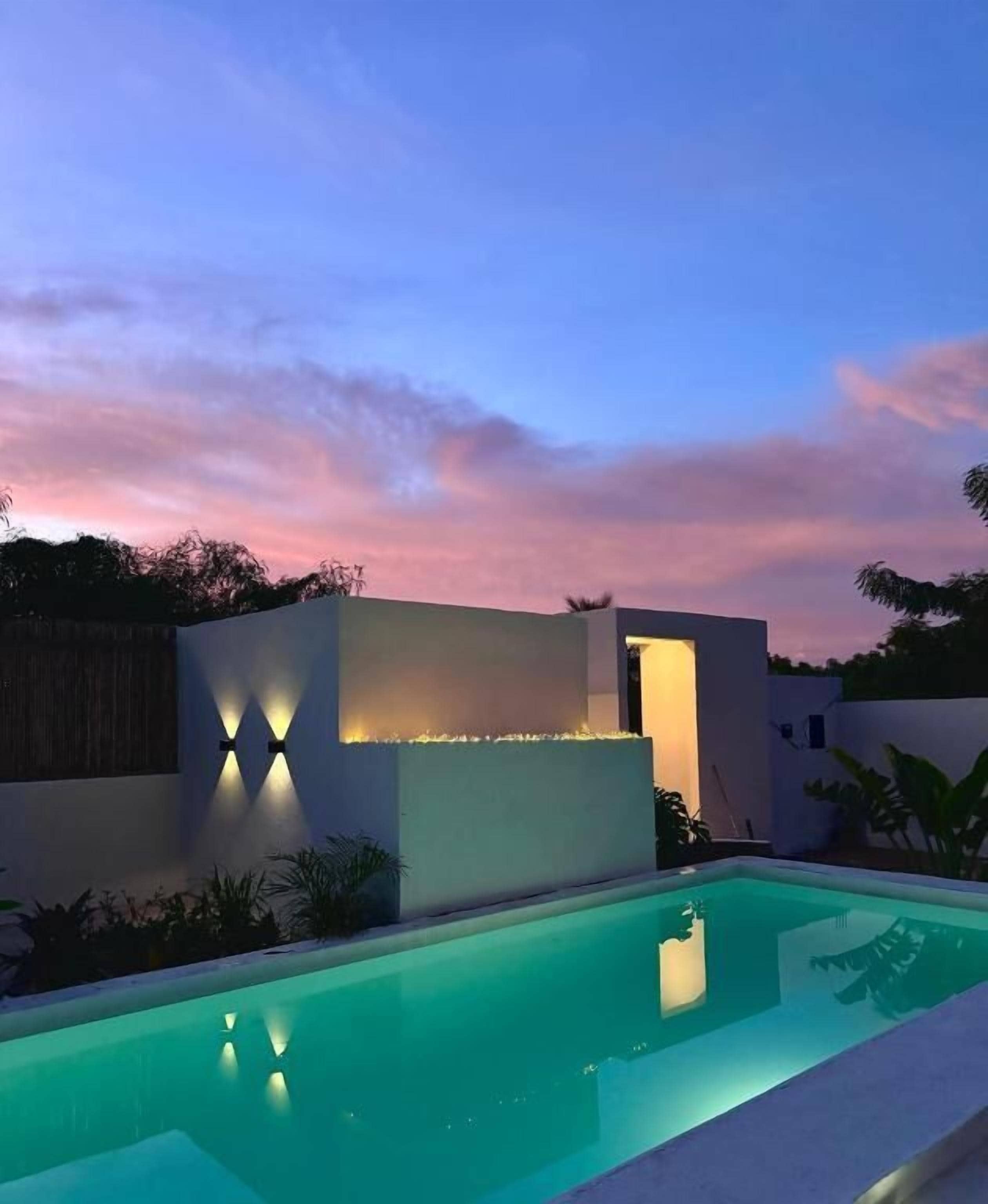 A modern outdoor pool area with a clear pool, a low wall with integrated lighting, and a sunset sky.