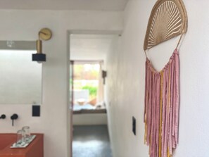 Un pasillo con pared blanca, un reloj, un ventilador y una puerta que da a un espacio luminoso.