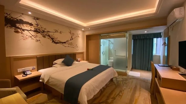 A hotel room with a bed, bedside table, chair, desk, and a bathroom visible through a glass door.