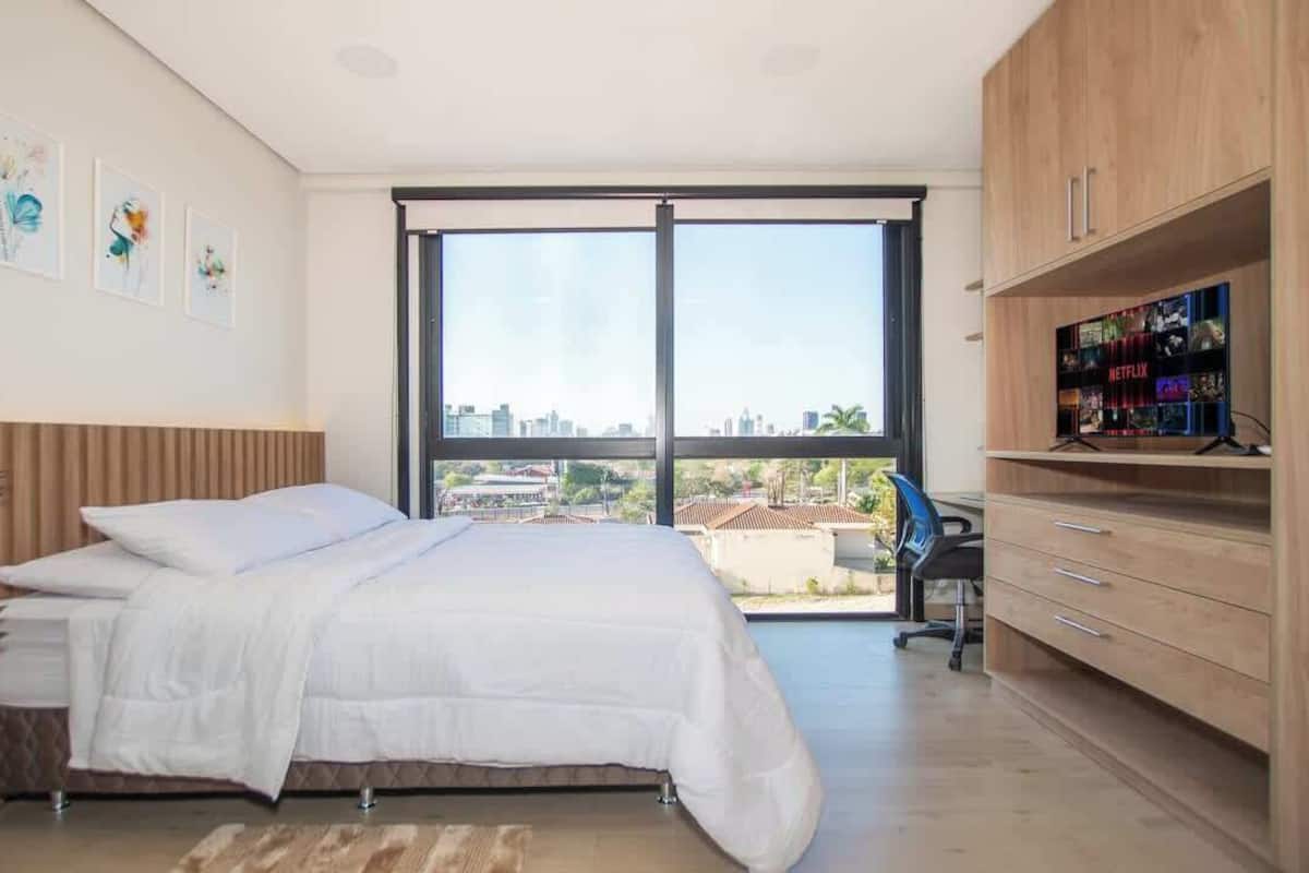 Un dormitorio con cama, escritorio, silla, televisor y un ventanal con vista a la ciudad.