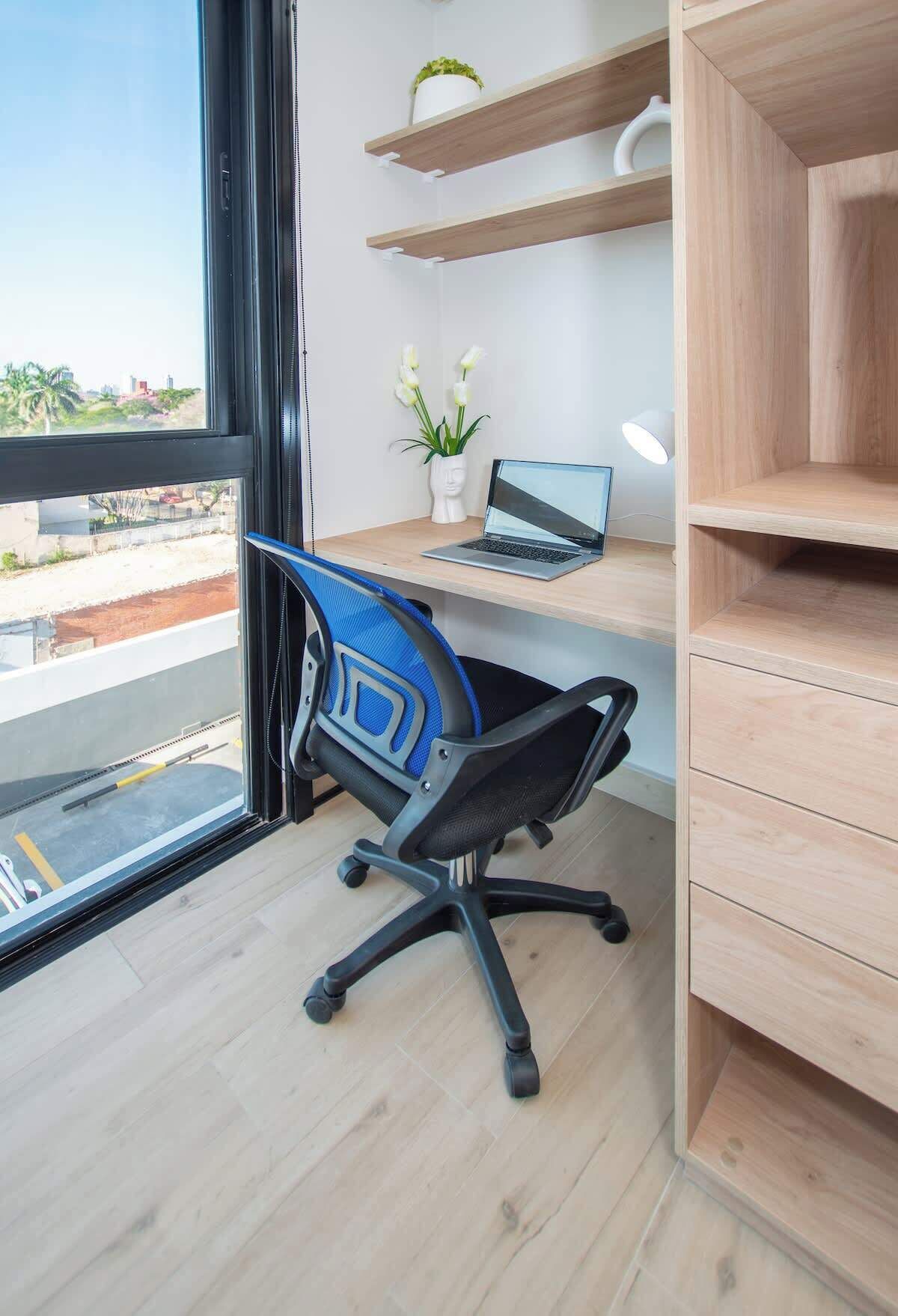 A modern office space with a desk, chair, laptop, shelves, and a view through a window.