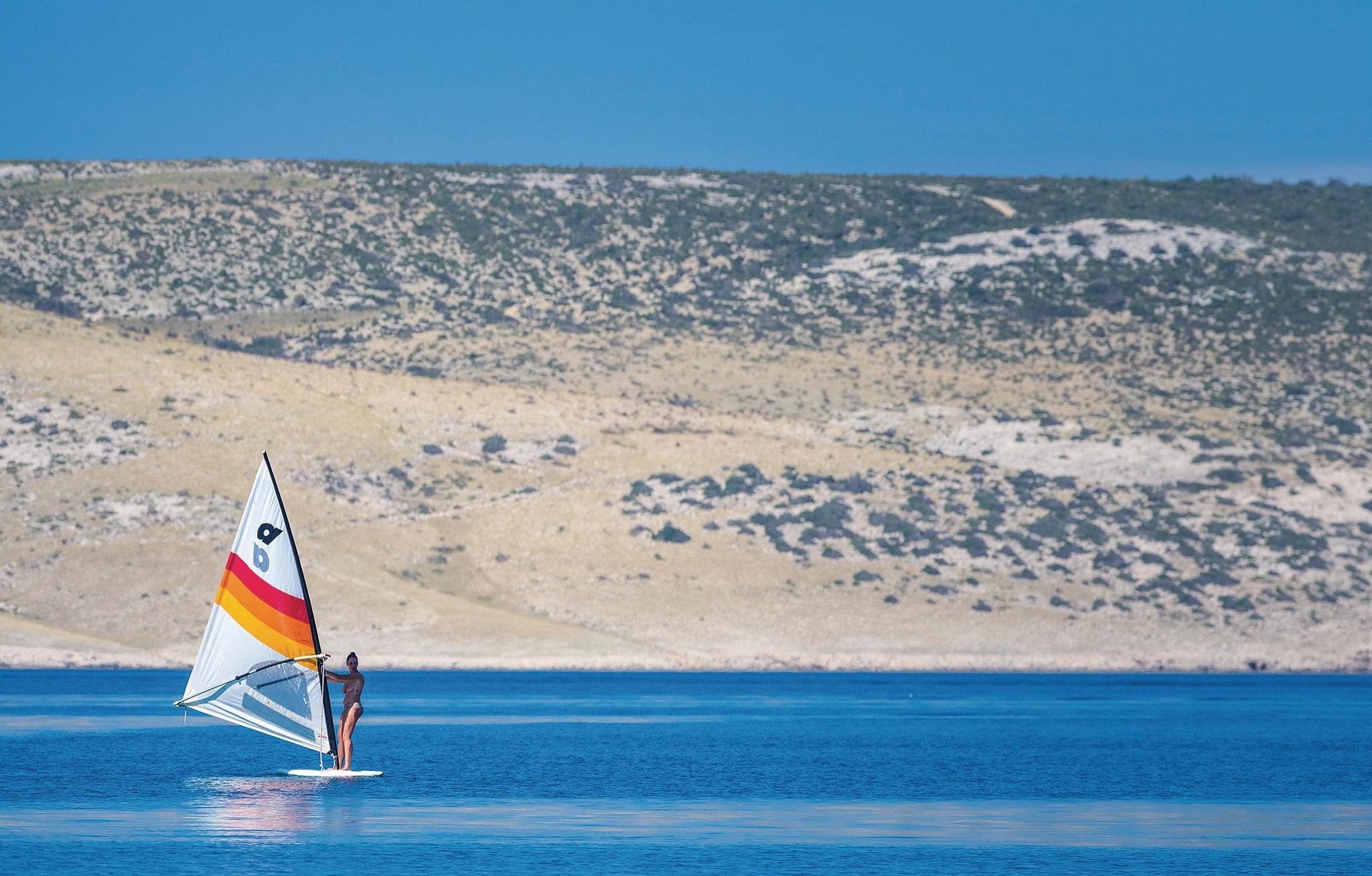 A person windsurfing on a clear day with a sandy beach and a hilly landscape in the background.