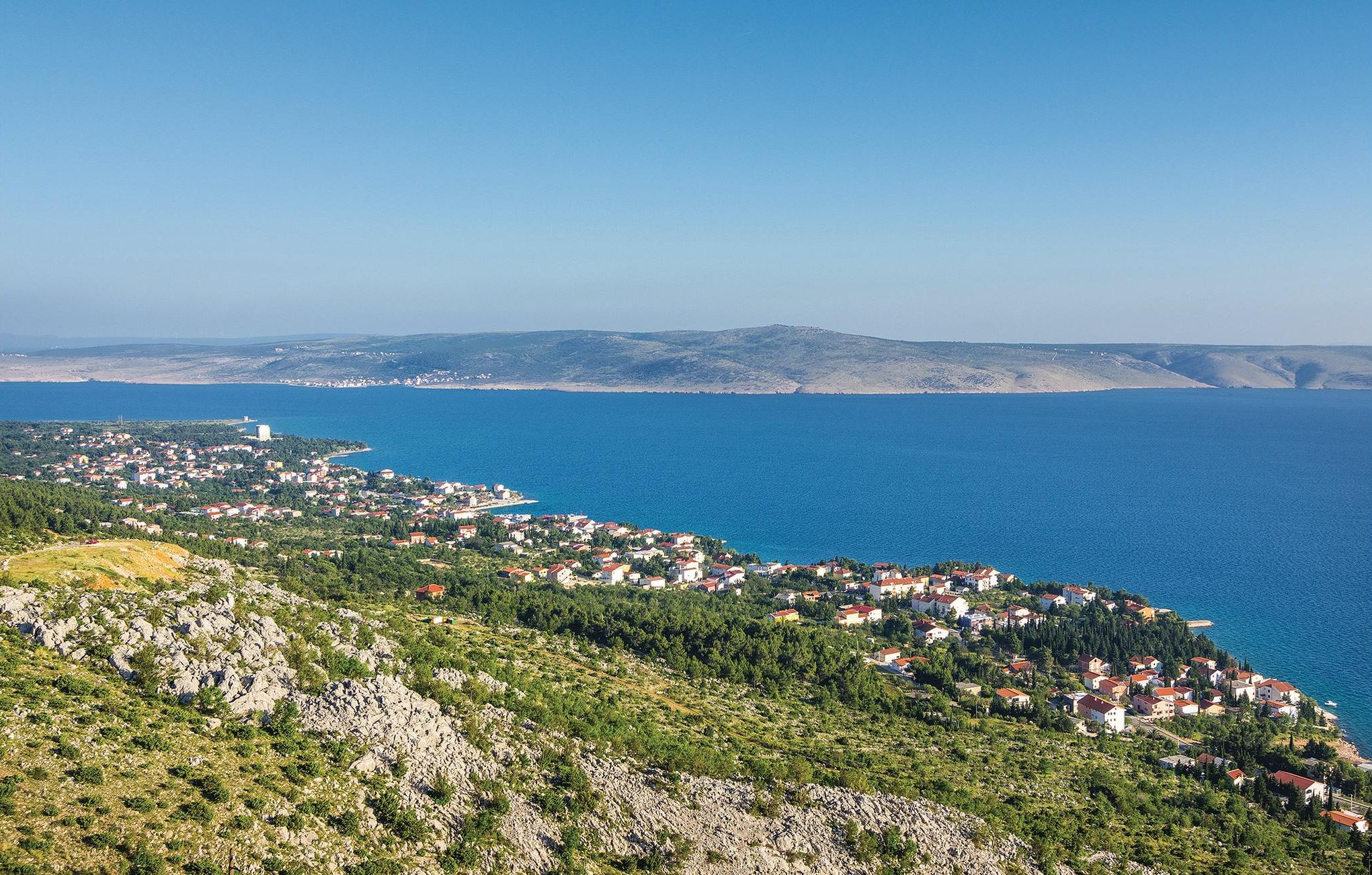 A coastal town with buildings along the shoreline, a clear blue sea, and hilly terrain in the background.
