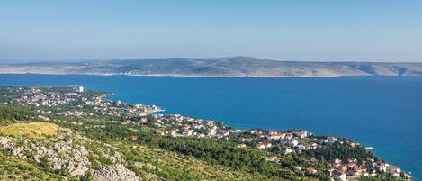 A coastal town with buildings along the shoreline, a clear blue sea, and hilly terrain in the background.